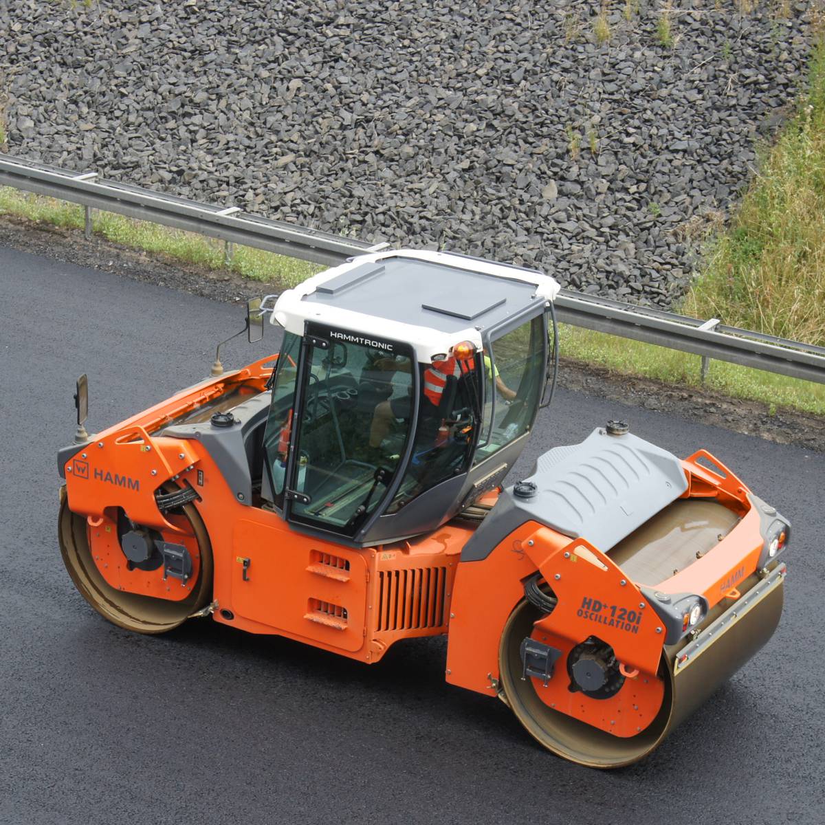 A HAMM HD+ 120i OV tandem roller compacting the surface course of a busy highway